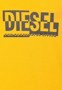 Camiseta de algodón amarilla con texto en negrita y gris oscuro "DIESEL" y "PARA UNA VIDA EXITOSA" en una fuente más pequeña debajo. Textura suave, diseño de cuello redondo.