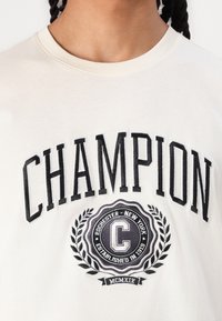Kretrainas kokvilnas T-krekls ar melno "CHAMPION" tekstu un apli logotipu, kas ietver lauru lapas un dibināšanas gadu. Apaļš kakla izgriezums, īsas piedurknes.