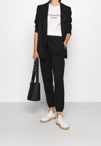 Kvinna som bär en svart kavaj och joggers, vit t-shirt med texten "Weekend MaxMara", vita sneakers och håller en svart bucket bag.