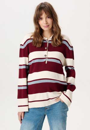 Vrouw met schouderlang bruin haar, gekleed in een bordeauxrode, witte en lichtblauw gestreepte longsleeve en blauwe spijkerbroek, poserend met één hand in de zak.