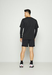 Μαύρο κοντομάνικο μπλουζάκι με πλέξη από διχτυωτό υλικό και μακρύτερα μαύρα μανίκια, συνδυασμένο με μαύρες βερμούδες και λευκά αθλητικά παπούτσια με κόκκινες λεπτομέρειες.