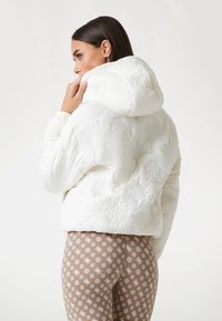 White quilted puffer jacket with a large hood and textured floral pattern, paired with light brown leggings featuring white geometric designs.