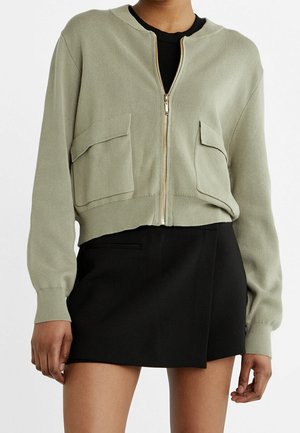 Femme portant un cardigan vert clair à fermeture éclair avec de grandes poches et une mini-jupe noire.