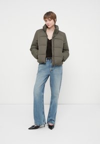 Ung kvinde i grøn pufferjakke, sort top, lyseblå brede jeans og sorte spidse hæle står imod en hvid baggrund.