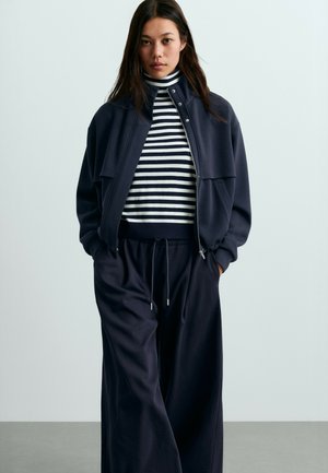 Jeune femme portant une veste bleu marine, un pantalon noir large et un pull à col roulé rayé noir et blanc, debout les mains dans les poches.