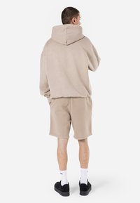 Beige oversized hættefyn med en stor hætte, blød tekstur og sænkede skuldre, parret med beige shorts og sorte sneakers.