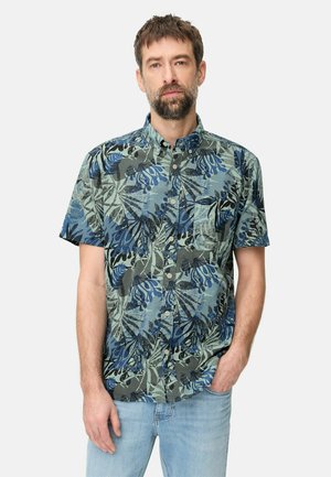 Man met baard die een blauwgroen overhemd met korte mouwen en abstracte bladprint draagt, gecombineerd met lichtblauwe spijkerbroek, hand in de zak.