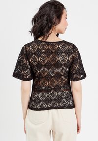 Top en dentelle noire avec un motif en losange, manches courtes et encolure ronde. Le tissu transparent met en valeur la peau en dessous.