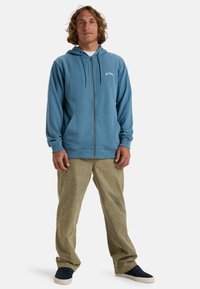 Veste à capuche zippée bleu avec cordons de serrage, poches avant et logo blanc. Assortie à un pantalon vert clair et des baskets noires. Look décontracté.