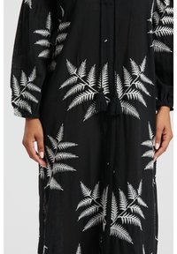 Czarna tkanina odzieżowa z białym haftem w kształcie paproci, z dekoltem ze sznurkiem i frędzlami. Luźny krój z długimi rękawami i bocznymi rozcięciami.