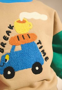Bēšas krāsas džemperis ar ziliem piedurknēm, ar teksturētu izšuvumu, kurā attēlots zils kravas auto, dzeltena krūze un uzraksts "BREAK TIME" melnā krāsā.