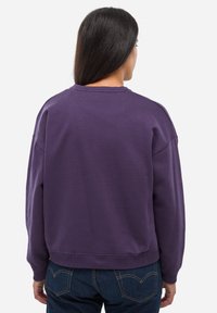 Fioletowy bawełniany sweatshirt z okrągłym dekoltem i opadłymi ramionami, zestawiony z ciemnymi dżinsami. Tkanina jest teksturowana i casualowa.