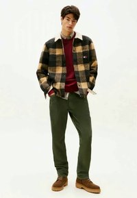 Plaid wool jacket in black and tan, red sweater, green pants, brown boots. Layered with a checkered shirt collar. Simple, casual design.