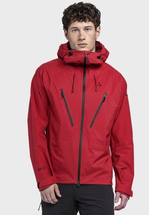 Junger Mann trägt eine rote wasserdichte Jacke mit Kapuze, zwei vertikalen, mit Reißverschluss versehenen Brusttaschen und schwarze Hose, steht vor neutralem Hintergrund.