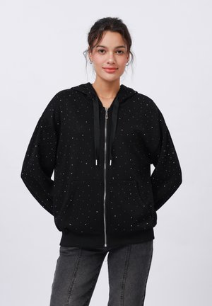 Sudadera negra con cremallera, hecha de un tejido suave, con tachuelas plateadas en un patrón disperso. Incluye capucha con cordón y bolsillos frontales.