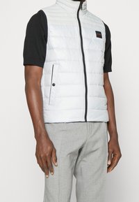 Ljusblå quiltad väst med hög krage, dragkedja fram och svarta detaljer; bärs över en kortärmad svart skjorta, ihop med grå byxor.