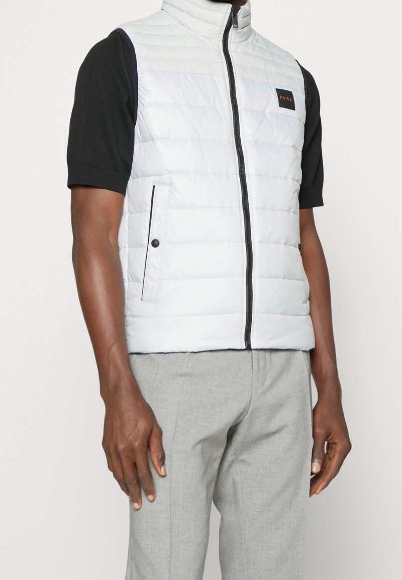 Ljusblå quiltad väst med hög krage, dragkedja fram och svarta detaljer; bärs över en kortärmad svart skjorta, ihop med grå byxor.