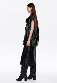 Melnas ādas veste ar īsām piedurknēm, apvienota ar saskaņojošām garām svārkiem un zābakiem. Gluda tekstūra un mirdzoša apdare visā virsmas laukumā.