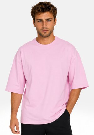 Giovane uomo con capelli castano chiaro e occhi azzurri, indossa una maglietta rosa larga e pantaloni neri, in piedi con una mano in tasca.