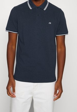 Mann trägt ein dunkelblaues Poloshirt mit weißen Streifen am Kragen und an den Ärmeln, kombiniert mit weißen Hosen.