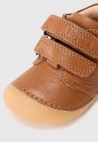 Scarpa per bambini in pelle marrone con due strappi in velcro, punta arrotondata e suola in gomma beige flessibile. Dettagli cuciti in vista.