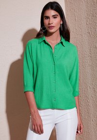 Camicia verde brillante con tessuto strutturato, maniche arrotolabili e vestibilità rilassata. Bottoni lisci sul davanti arricchiscono il design.