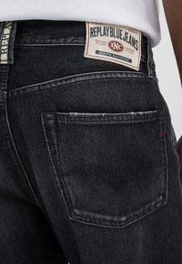 Black denim jeans featuring a visible back pocket, classic stitching, and a branded label at the waistband. Textured fabric with a faded finish.
