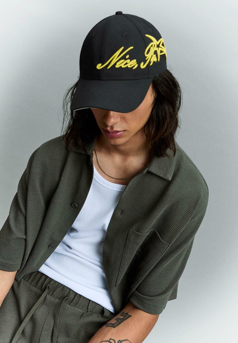 Personne portant une casquette noire avec le texte jaune "Nice, I'm" et une étoile, une chemise texturée olive à manches courtes, un débardeur blanc et un pantalon assorti, regardant vers le bas.