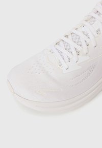 Chaussure de sport blanche avec une tige texturée, des lacets fins et une semelle en caoutchouc lisse. Dispose d'aérations pour la respirabilité et d'accents de design minimalistes.