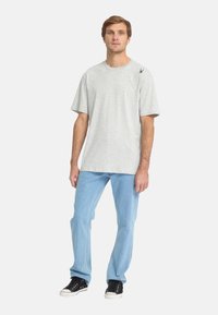Lichtgrijs T-shirt met korte mouwen, een comfortabele pasvorm en een klein zwart spinnenlogo op de schouder, gecombineerd met lichtblauwe jeans en zwarte sneakers.