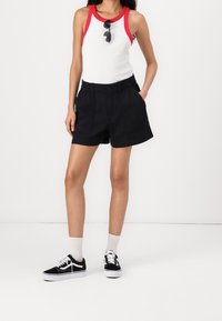 Černé šortky s předními kapsami, nošené s bílým tílkem s červeným límečkem, doplněné černými teniskami a kotníkovými ponožkami.