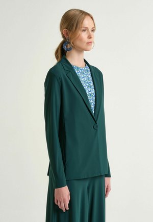 Donna con blazer verde e gonna, con top a motivo blu e verde, che indossa grandi orecchini circolari blu, in piedi davanti a uno sfondo chiaro uniforme.