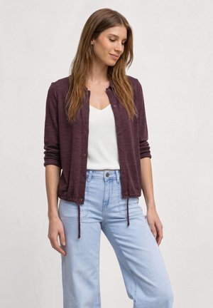 Jeune femme portant un cardigan bordeaux boutonné, un haut blanc et un jean bleu clair, debout devant un fond clair uni, regardant vers le bas.