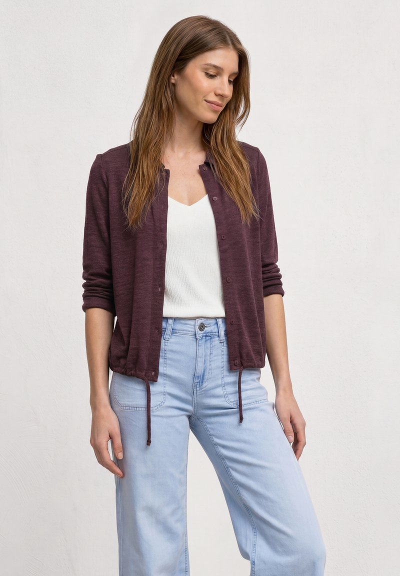 Jeune femme portant un cardigan bordeaux boutonné, un haut blanc et un jean bleu clair, debout devant un fond clair uni, regardant vers le bas.