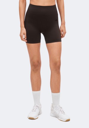 Osoba u crnim sportskim kratkim hlačama s visokim strukom, bijelim čarapama i bijelim tenisicama za trčanje, stoji na svijetloj pozadini.