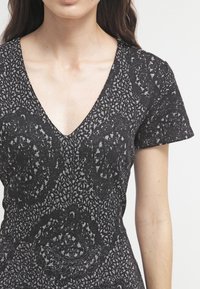 Frau trägt ein kurzärmliges schwarzes Kleid mit texturierten grauen runden und abstrakten Mustern, V-Ausschnitt und taillierter Passform.