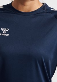 Námořnická modrá sportovní košile vyrobená z hladkého materiálu, s kulatým výstřihem a bílým logem na levé části hrudi, s bílými akcenty na ramenou.
