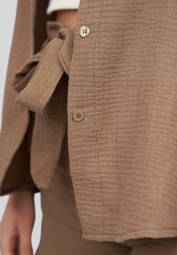 Cardigan in tessuto marrone con texture, chiuso da una cintura e bottoni piccoli. Presenta un motivo a righe verticali e un orlo pulito.
