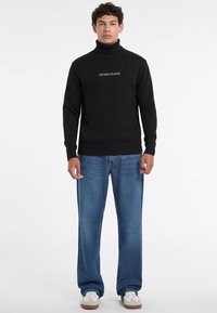 Maglione nero a dolcevita con polsini e orlo a costine, caratterizzato dal logo bianco "GUESS JEANS" sul petto, abbinato a jeans a gamba larga di colore blu chiaro.