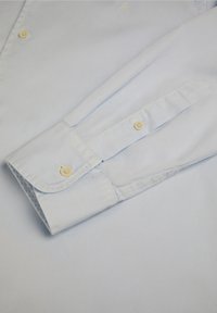Cămașă cu butoane de culoare albastru deschis, cu o textură netedă, având un manșon pliat cu închidere prin buton și un detaliu subtil de broderie pe piept.