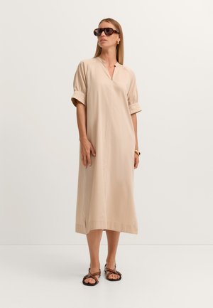 Frau trägt ein beiges Midikleid mit kurzen Ärmeln, dunkle Sonnenbrille, braune Sandalen, goldene Creolenohrringe und eine Armbanduhr, steht vor weißem Hintergrund.