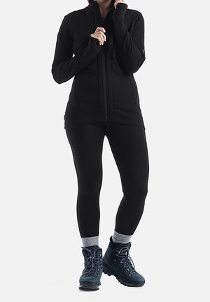 Person iført sort lynlåstrøje, sorte leggings, grå sokker og mørkeblå vandrestøvler, stående mod en ensfarvet baggrund.