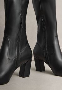 Svarta läderstövlar till knäna med slät yta, fyrkantiga klackar och sidoblixtlås. Har en elegant, åtsittande design för en strömlinjeformad look.