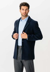 Mand justerer en navy blå blazer over en lyseblå stribet skjorte, iført grå bukser, stående mod en simpel hvid baggrund.