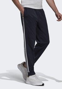 Marineblaue Jogginghose mit weißen Seitenstreifen, aus leichtem Material; verfügt über seitliche Taschen und eine lockere Passform. Kombiniert mit weißen Sneakers.
