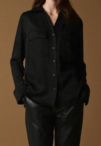 Černá košile s dlouhým rukávem a knoflíkovým zapínáním vpředu, dvěma náprsními kapsami a hladkou texturou, zkombinovaná s černými koženými kalhotami.