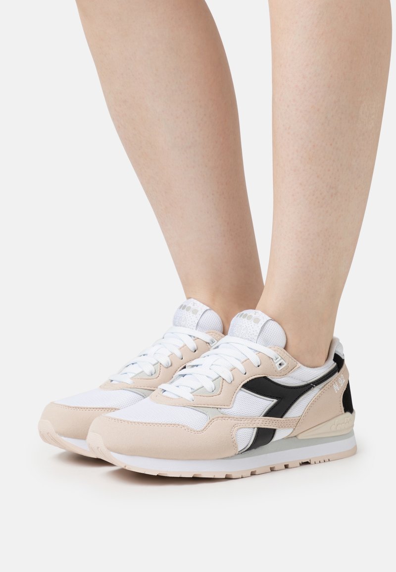 Beige and white sneakers with black accents, featuring a mesh and suede upper, lace-up closure, and cushioned sole. Textured detailing on the side.