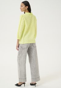 Top verde lime, semi-trasparente, con texture ondulata e scollatura a volant, abbinato a jeans grigi chiari a gamba larga e sandali neri con tacco.