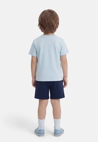 Παιδί που στέκεται με την πλάτη, φορώντας ανοιχτό μπλε μπλουζάκι, σκούρα μπλε σορτς, λευκές κάλτσες και ανοιχτό μπλε αθλητικά παπούτσια, με φόντο ένα απλό λευκό φόντο.