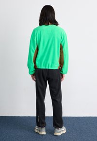 Grüner Fleece-Pullover mit braunen Seitenpanelen, schwarze Hose und graue Sneaker. Der Pullover und die Hose haben ein Logo, schlankes Design.